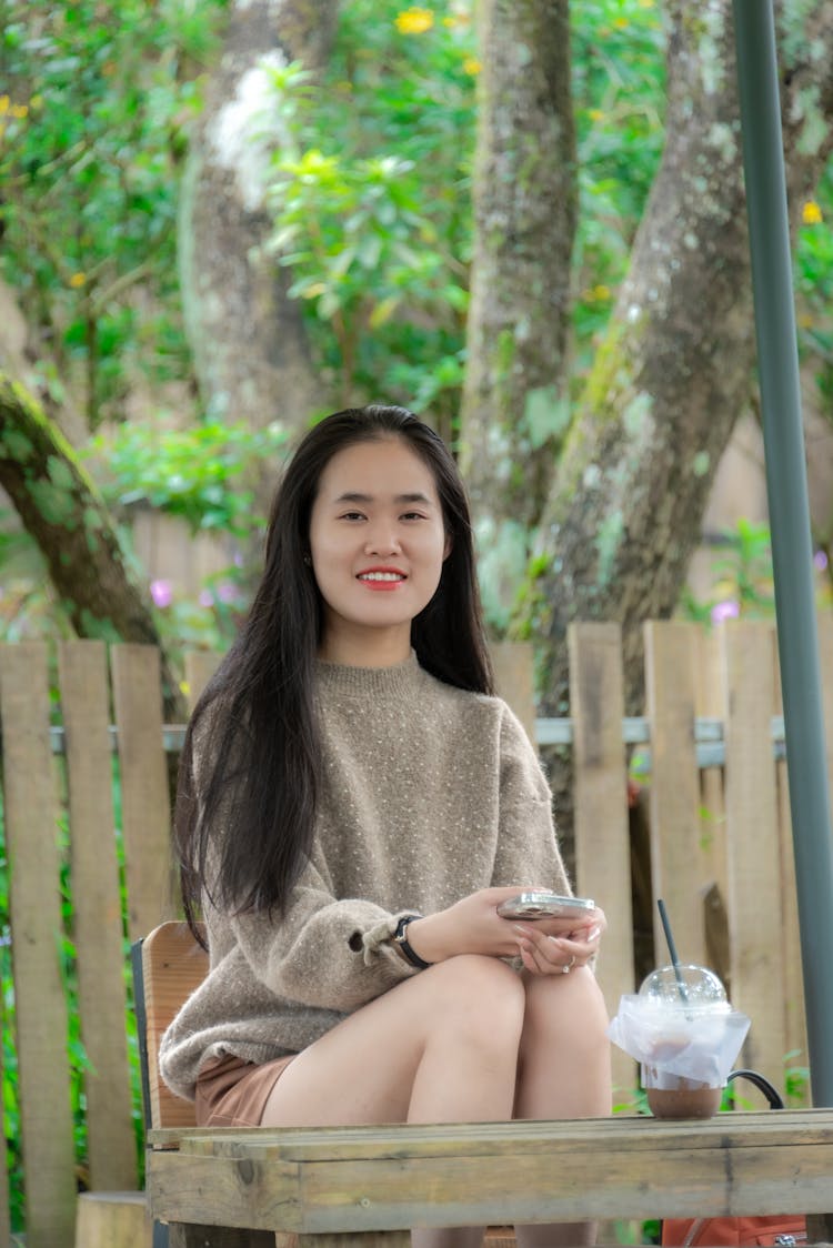 Young Asian Woman Sitting At Wooden Table In Summer Garden