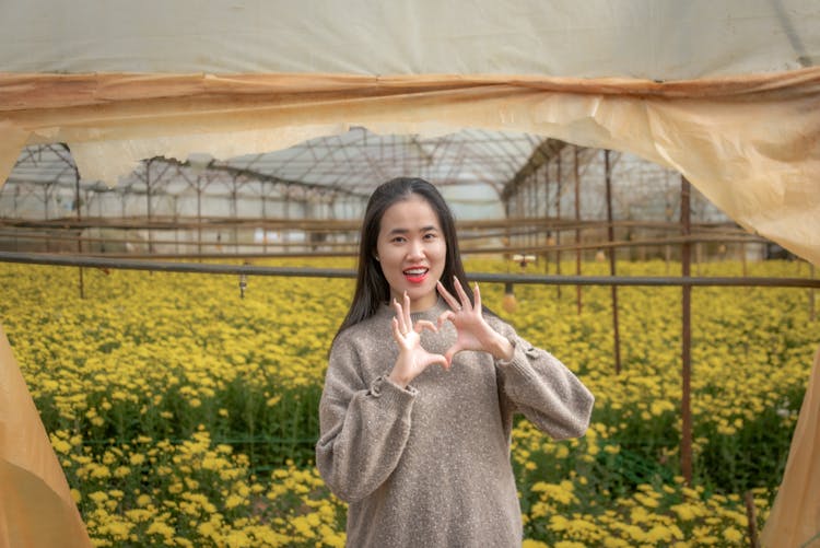 Smiling Asian Woman Showing Heart Hand Gesture In Greenhouse