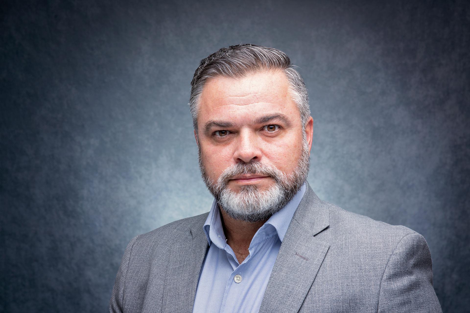 Studio portrait of a mature man with a beard and gray suit against a textured gray background.