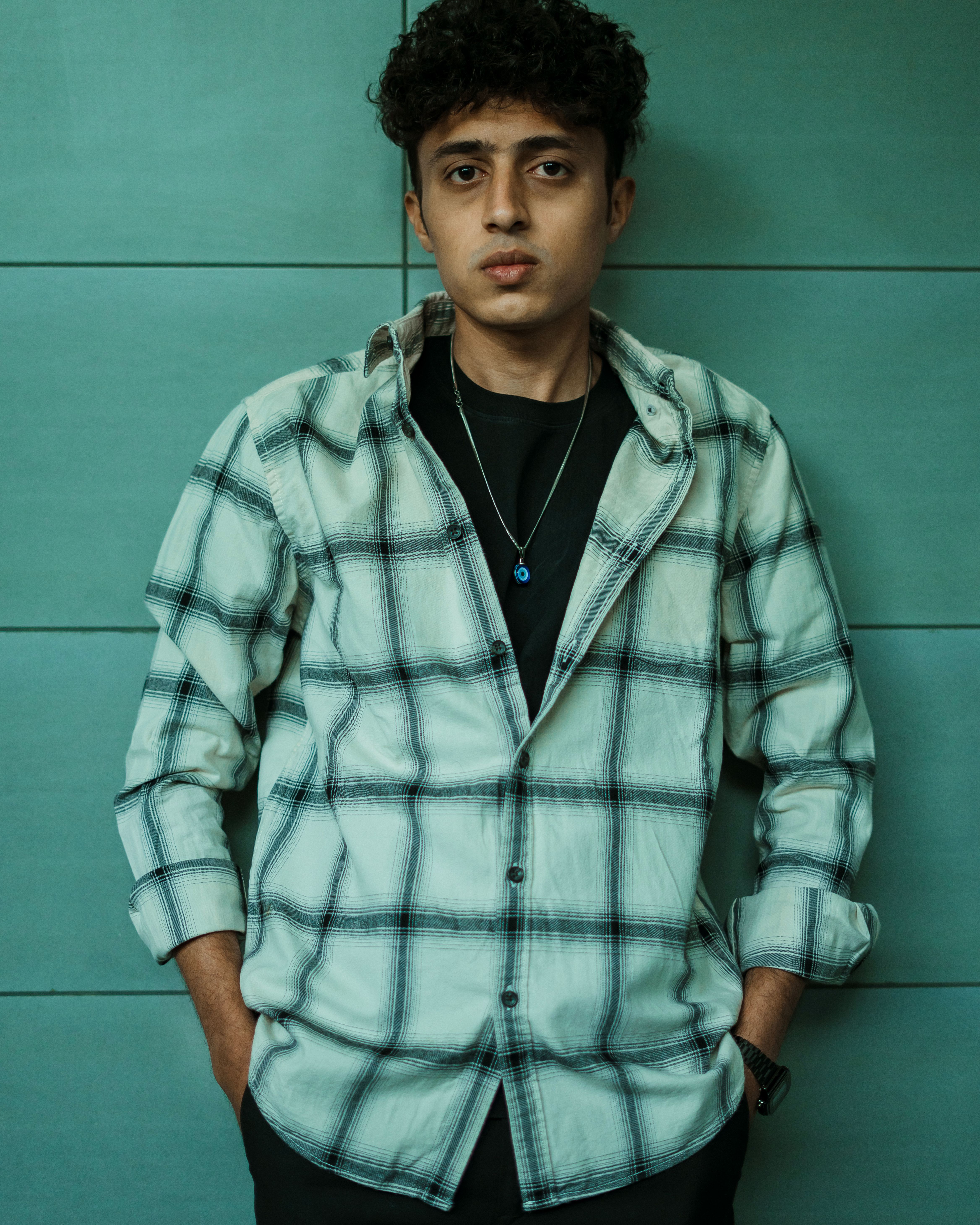 Young man standing confidently in checkered shirt with hands in pockets, showcasing modern fashion.