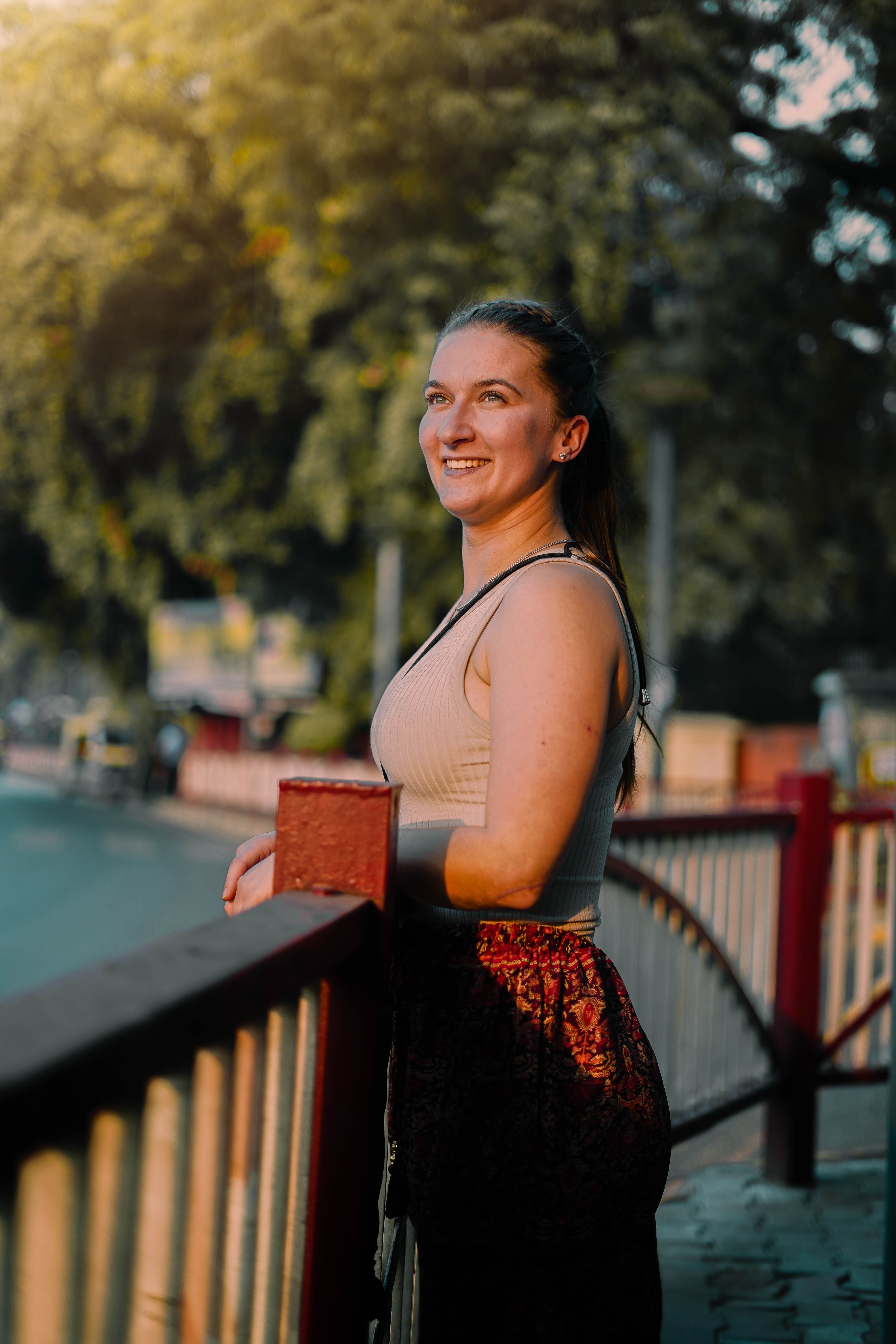 A woman smiles while leaning on a railing in a sunlit urban area.