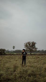 A solitary man in a t-shirt stands in a serene rural field with scattered trees under an overcast sky.