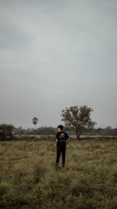 Free A solitary man in a t-shirt stands in a serene rural field with scattered trees under an overcast sky. Stock Photo
