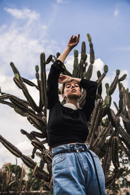Fashionable woman posing with arms raised in front of large cactus, blue sky background.