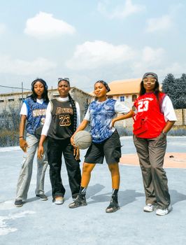 Four young women in sports jerseys pose confidently on an outdoor basketball court.