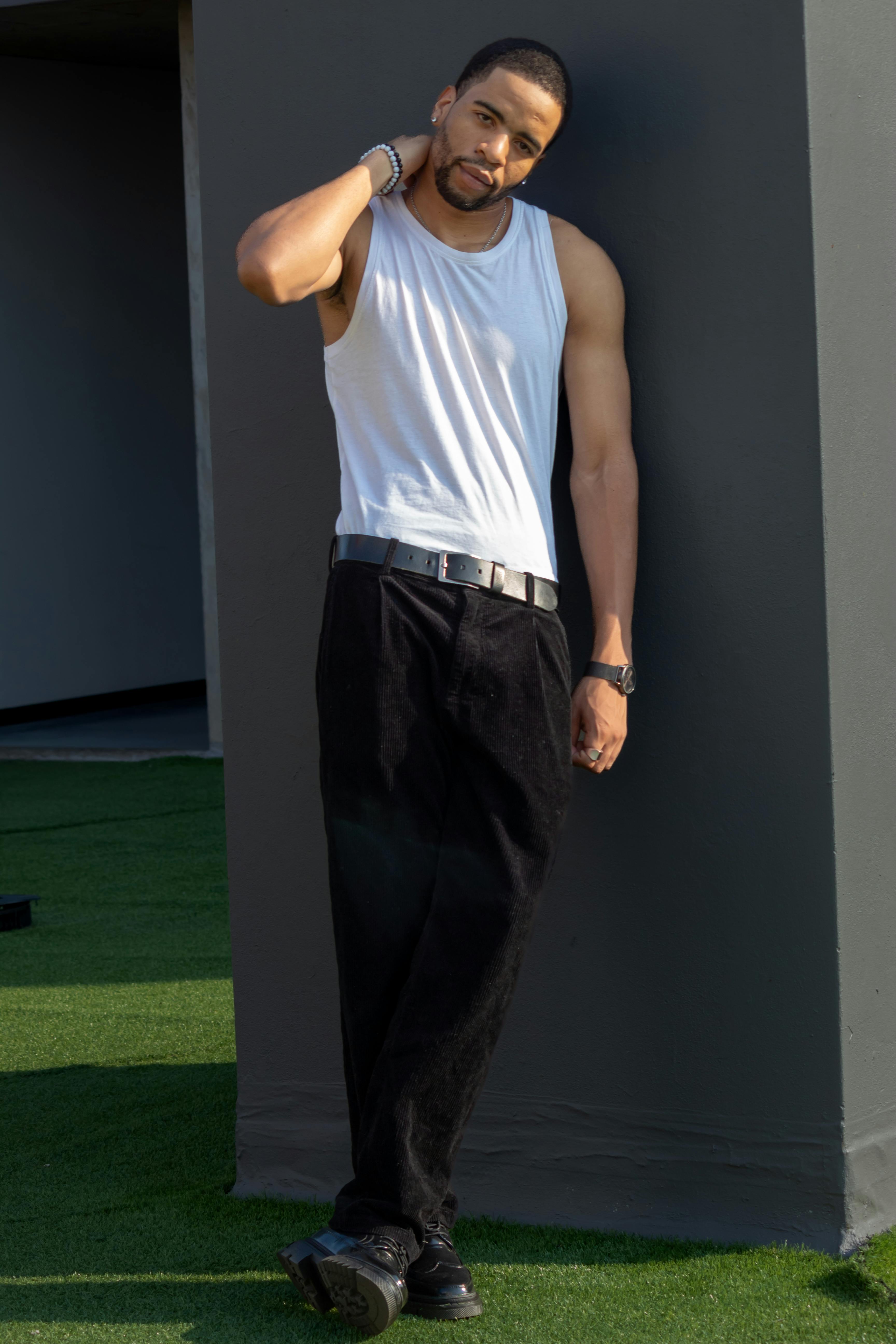 Stylish man in a white tank top and black pants poses confidently outdoors.