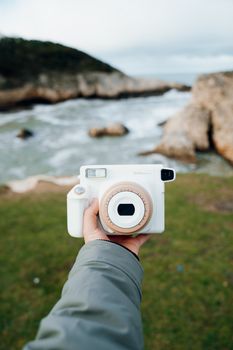 A person holds a retro camera with cliffs and ocean in the background, captured in a vertical shot.