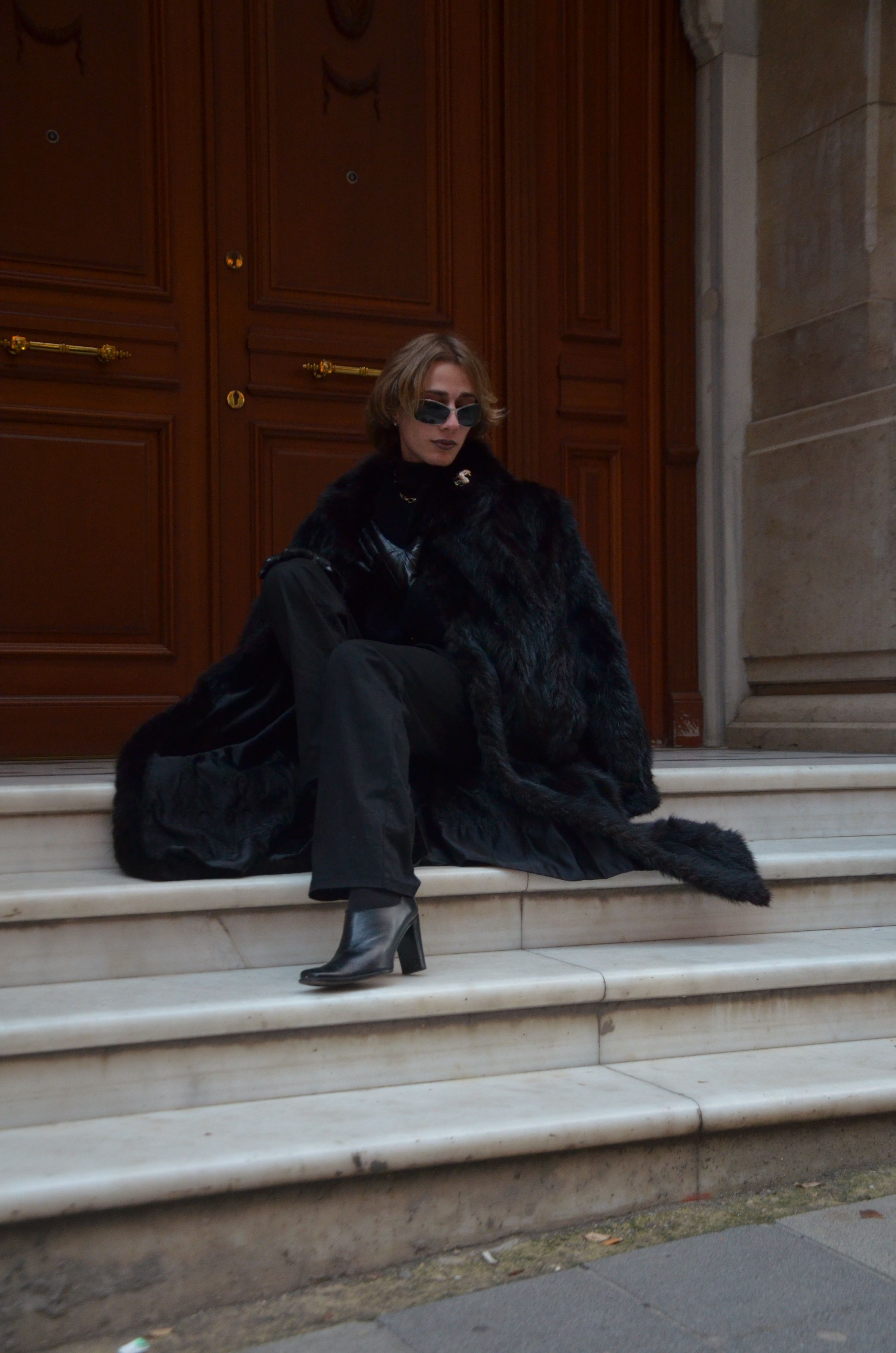Elegant woman in a black fur coat sitting on city steps, exuding confidence and style.