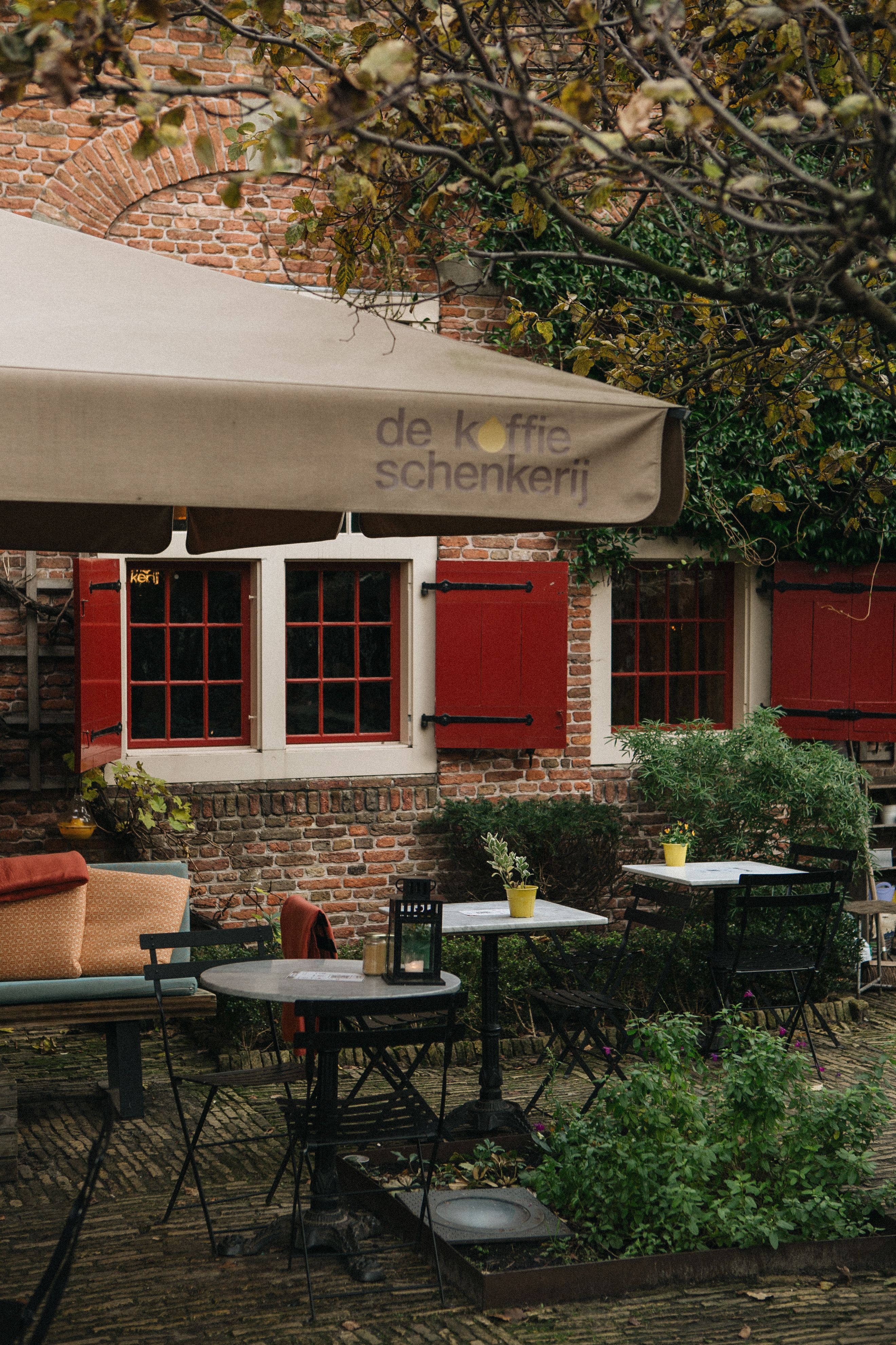Cozy urban café patio with empty tables, chairs, and lush greenery against a rustic brick wall.