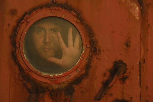 A Man Standing behind Rusty Door with a Circular Window 