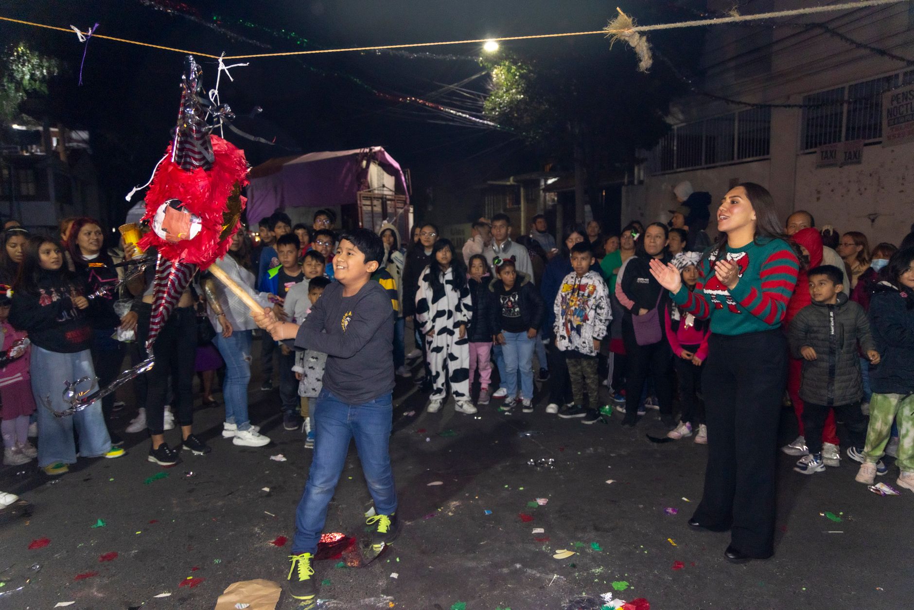 A vibrant street party in Coyoacán, Mexico, featuring a child hitting a piñata surrounded by a lively crowd at night.