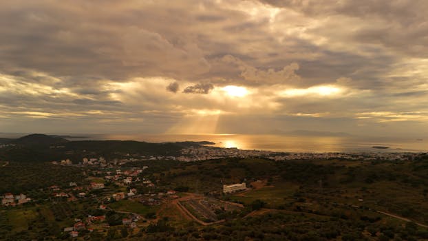 A breathtaking aerial view of Vari, Greece at sunrise with sun rays piercing through dramatic clouds over the sea.