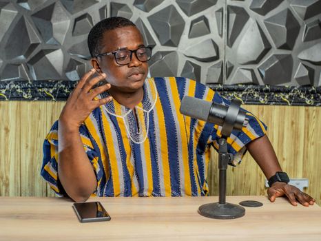 An adult man in a colorful Kente shirt recording a podcast. Indoor setting with microphone and smartphone.