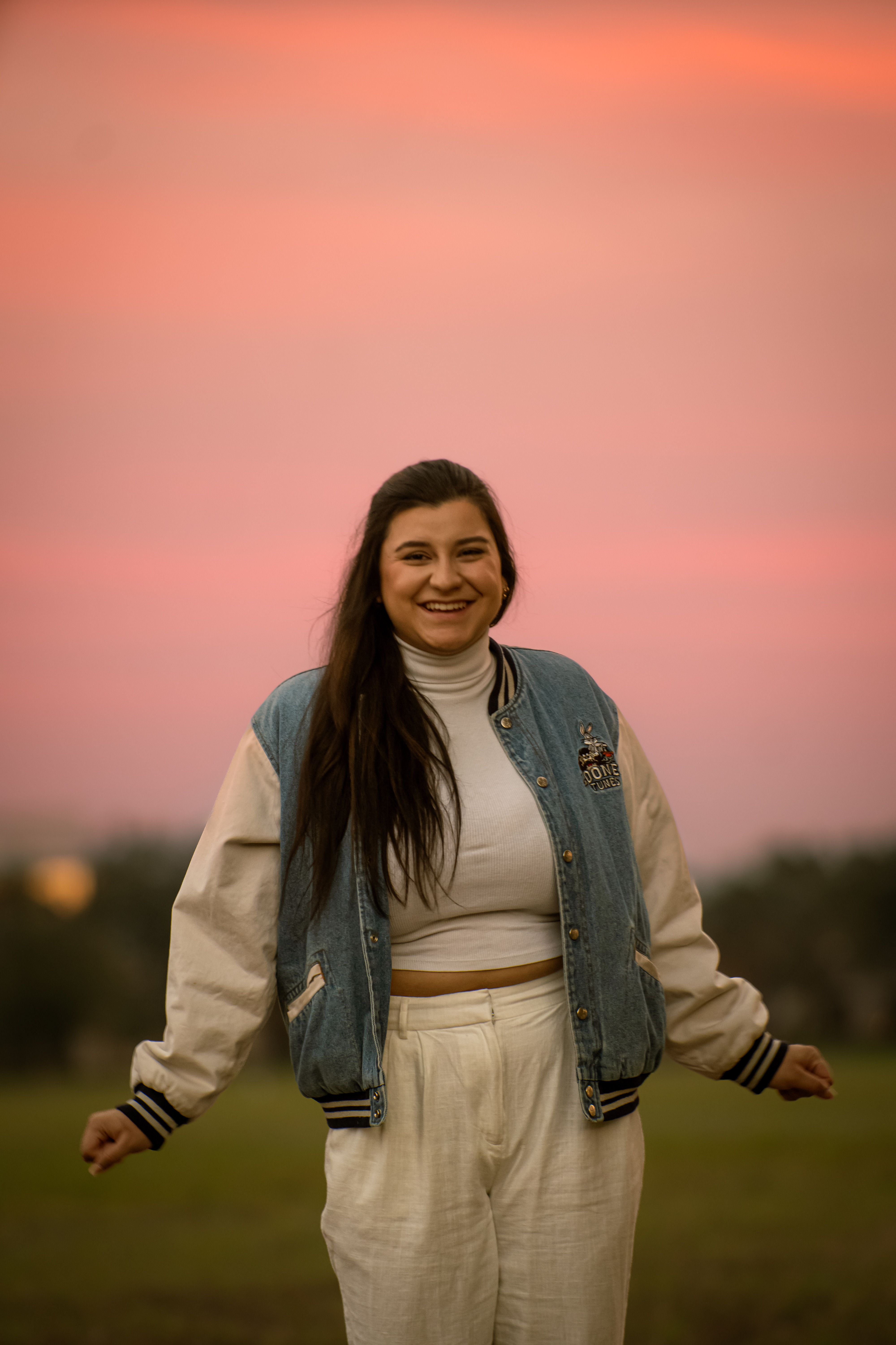 A joyful woman in a denim jacket smiling during a stunning sunset outdoors.
