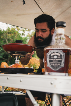 Bearded man preparing food outdoors with a camping stove and bottle. Captured in Natividade da Serra, Brazil.