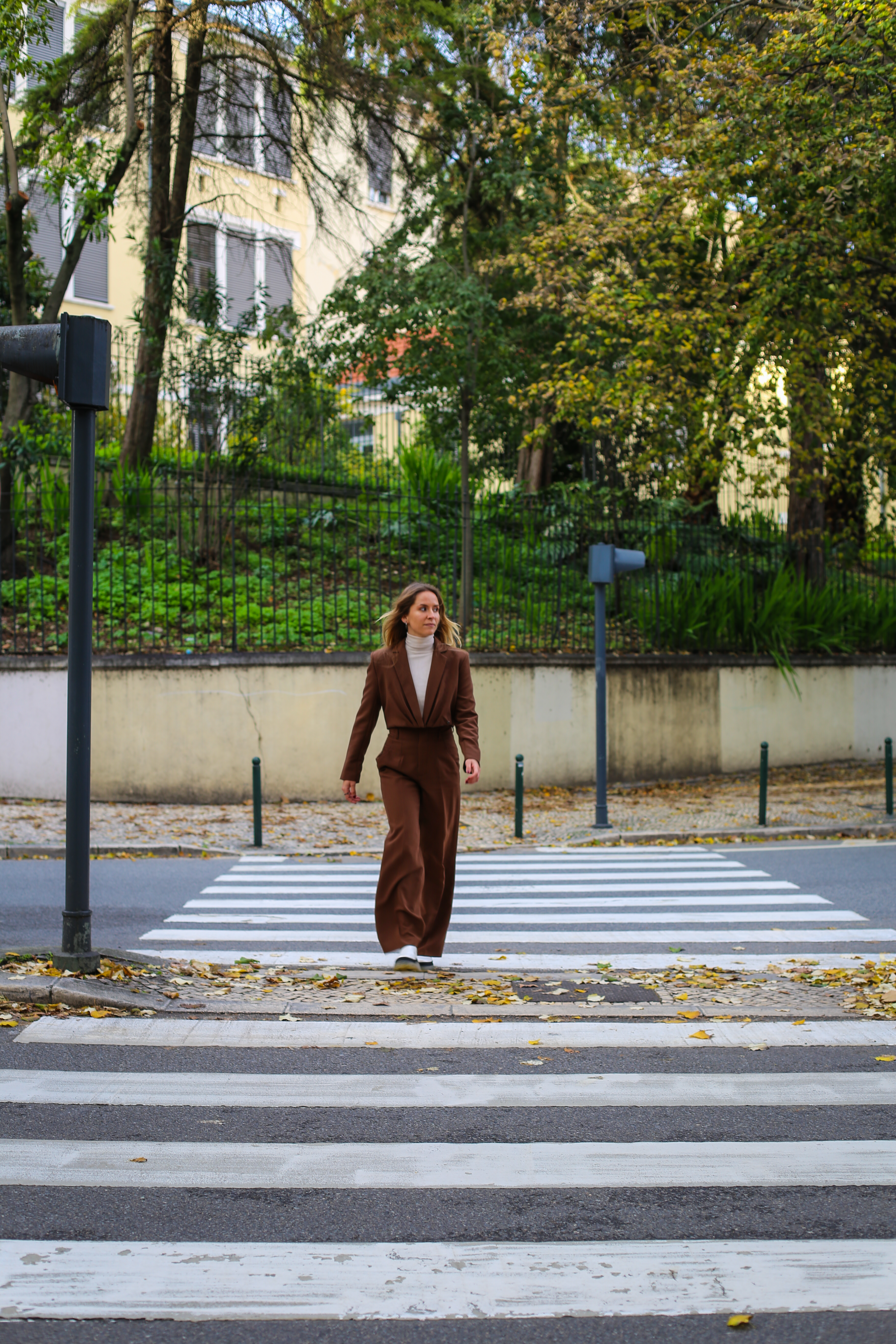 Woman in brown outfit walking on a city crosswalk surrounded by urban greenery in autumn.