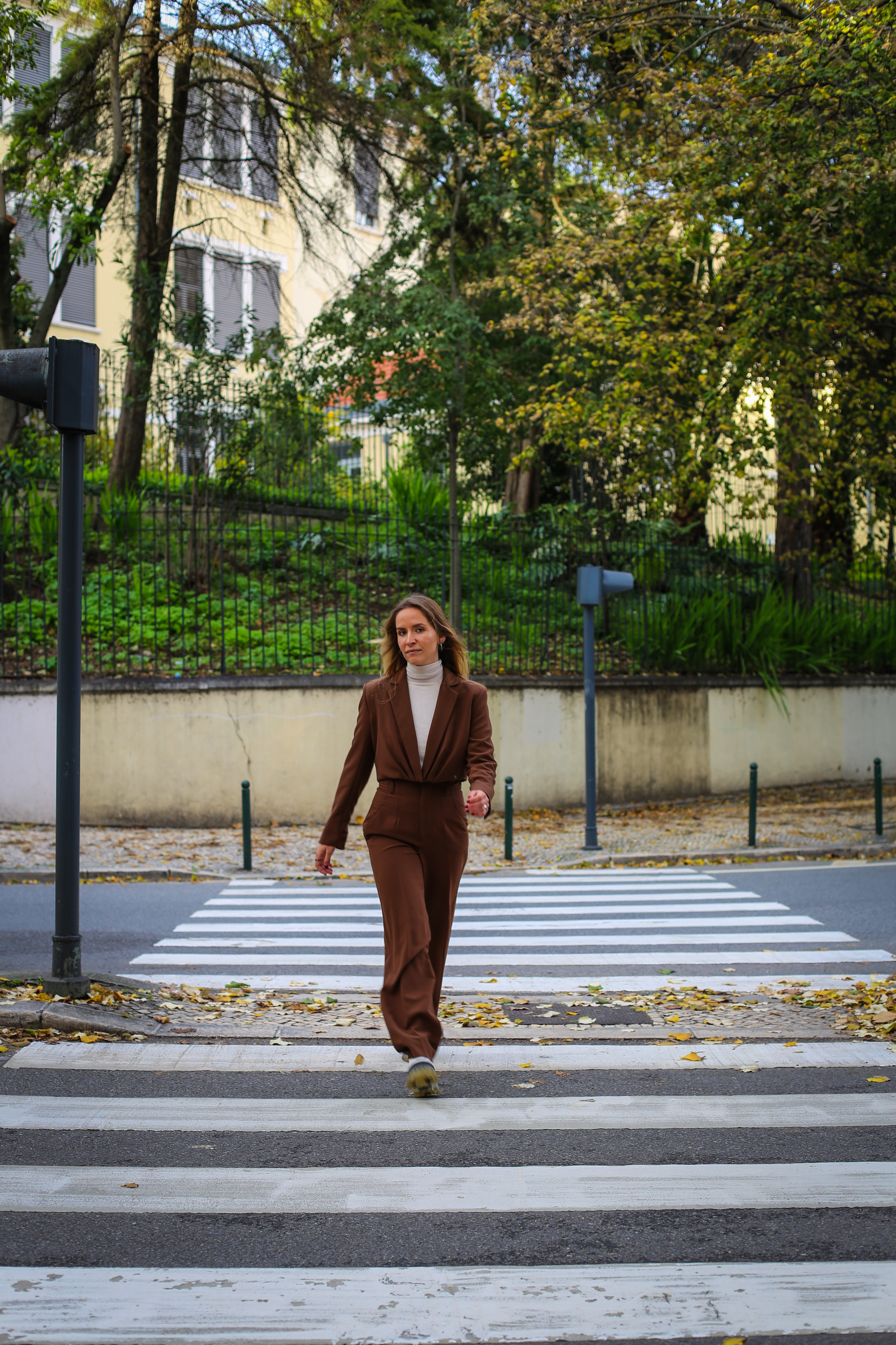 A woman in a stylish brown outfit walks across a city crosswalk surrounded by autumn foliage.