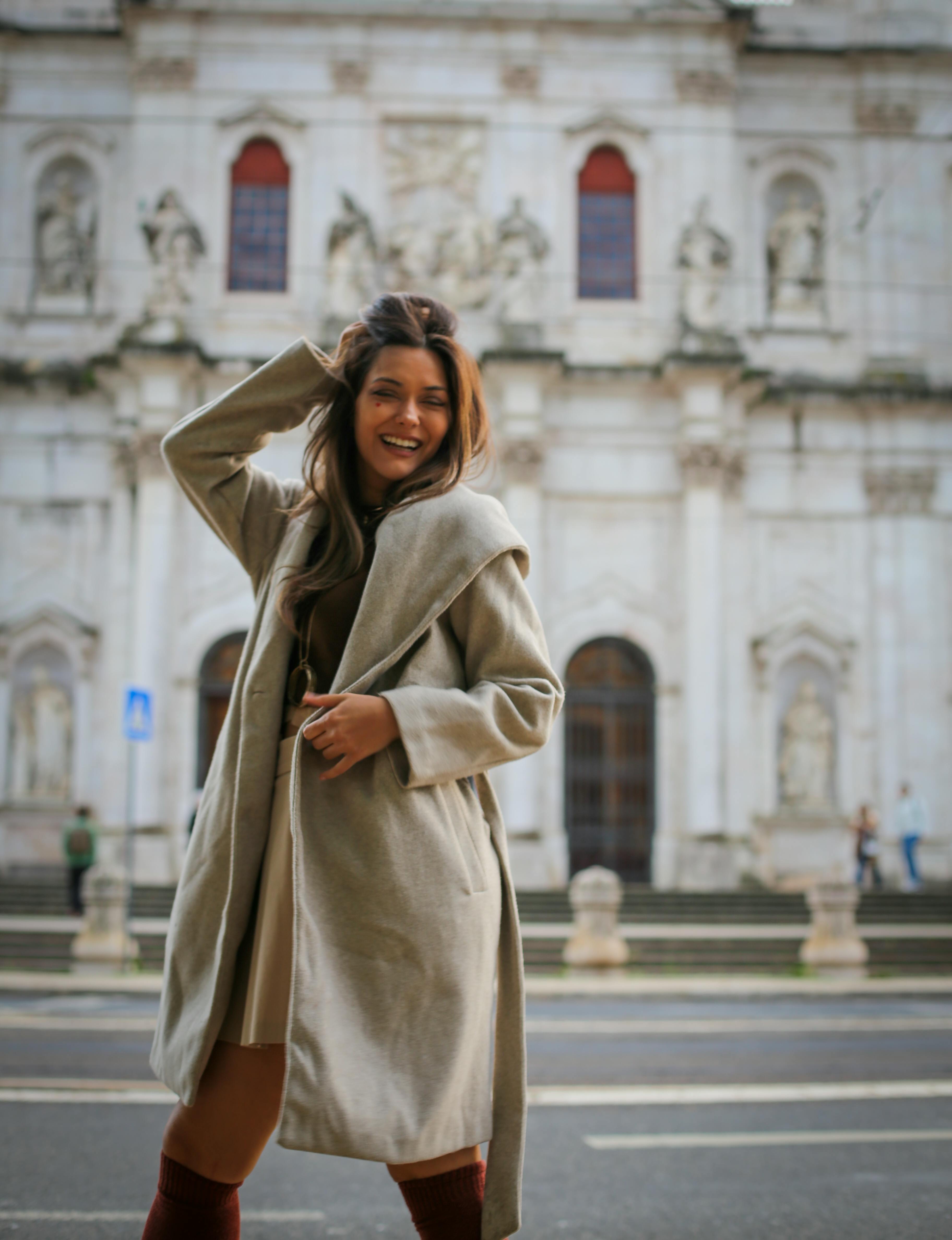 Young woman in a coat smiles playfully in front of an ornate building.