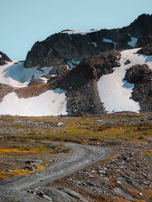 A dirt road leads through a scenic mountain landscape with snow-covered peaks under a clear sky.