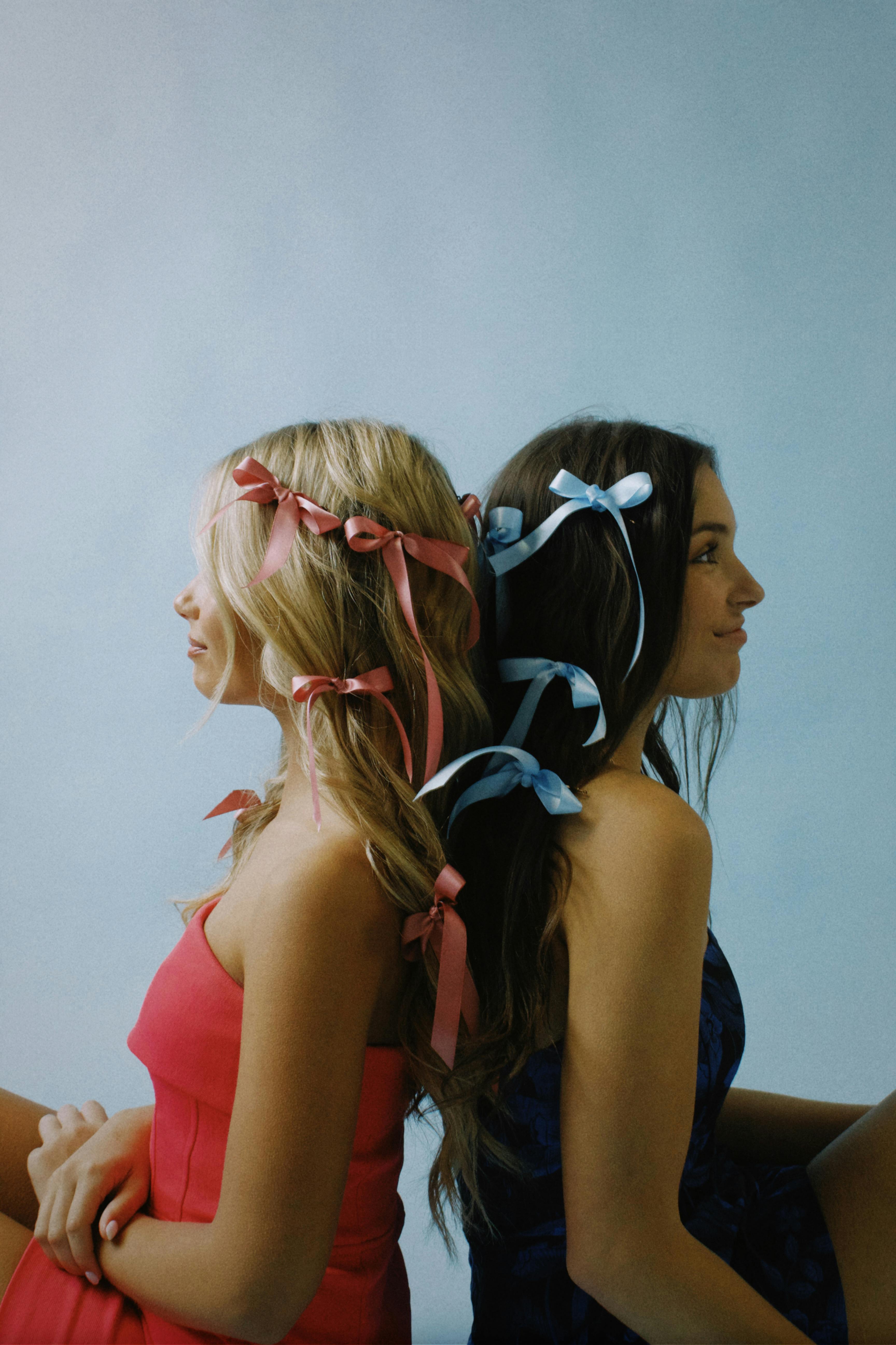 Two young women with colorful ribbons in their hair sitting back to back, exuding style and youth.