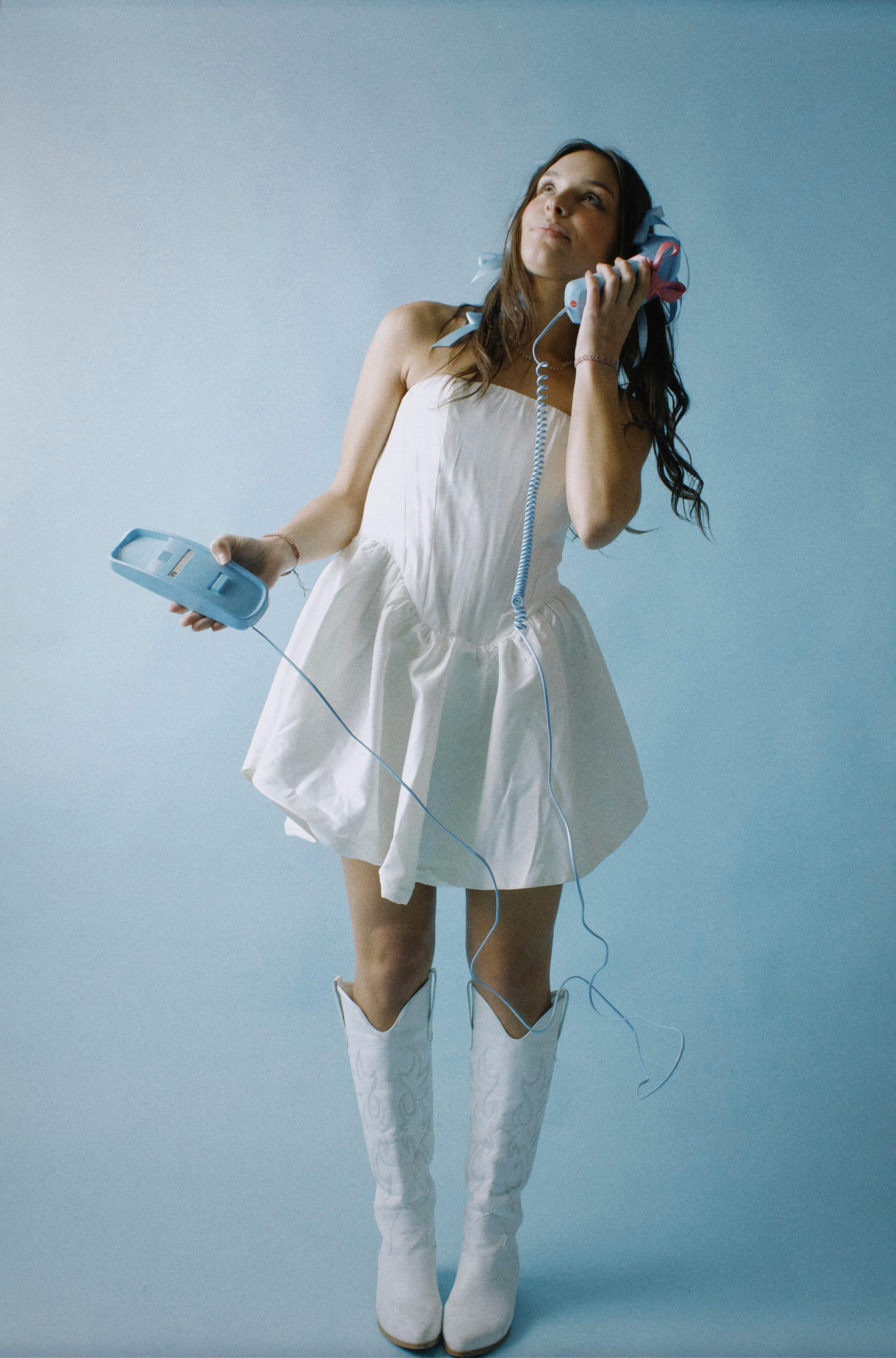 Woman in white dress and boots holds vintage telephone in a retro studio setup.