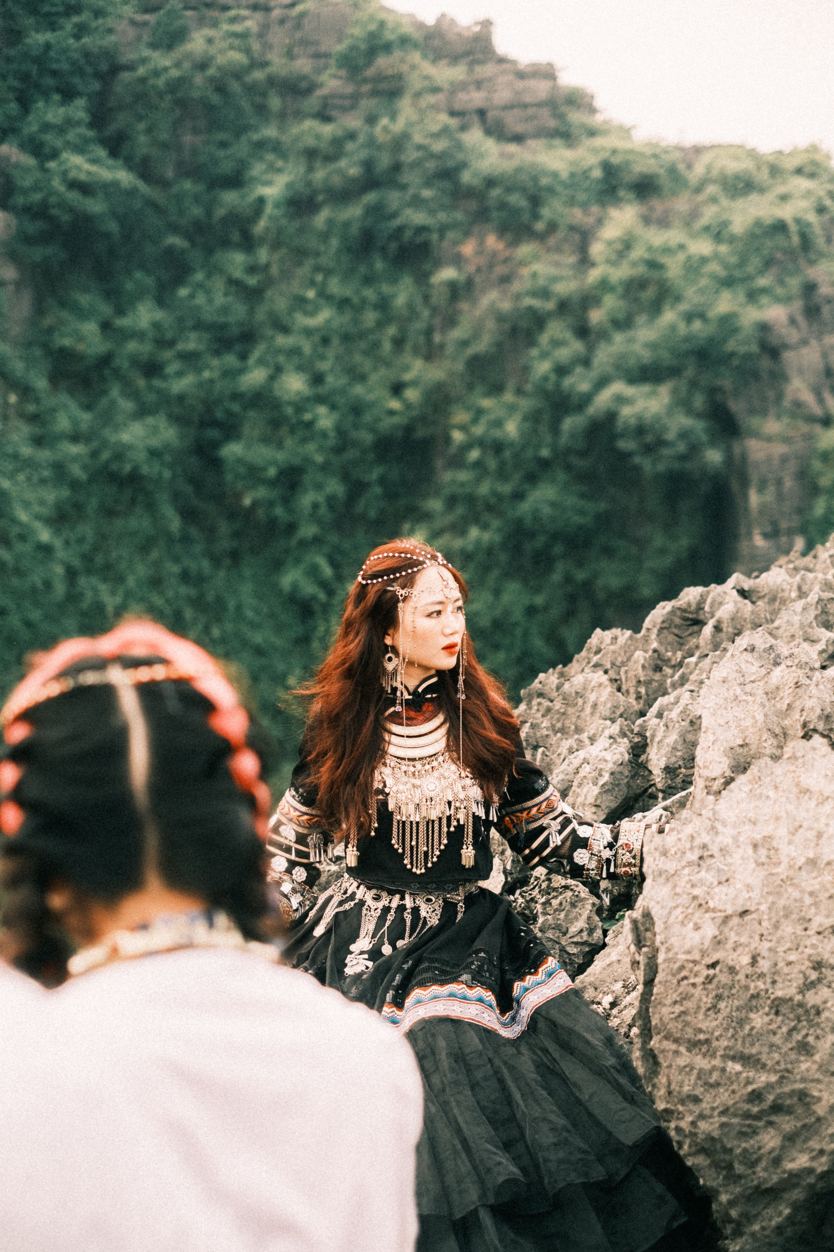 A woman in traditional Miao attire sits on a rocky mountain trail, capturing cultural elegance.