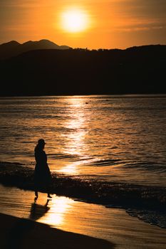 A stunning silhouette of a woman walking along the serene beach during sunset, reflecting golden hues.
