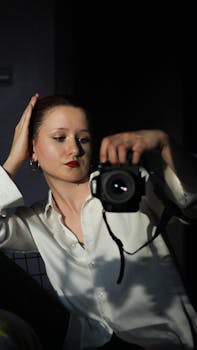 Young woman in a white shirt adjusting hair while holding a camera in low light.