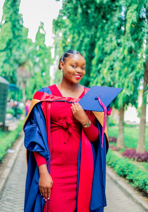 Smiling Student in Graduate Gown and Red Dress Holding a Mortarboard on a Walkway
