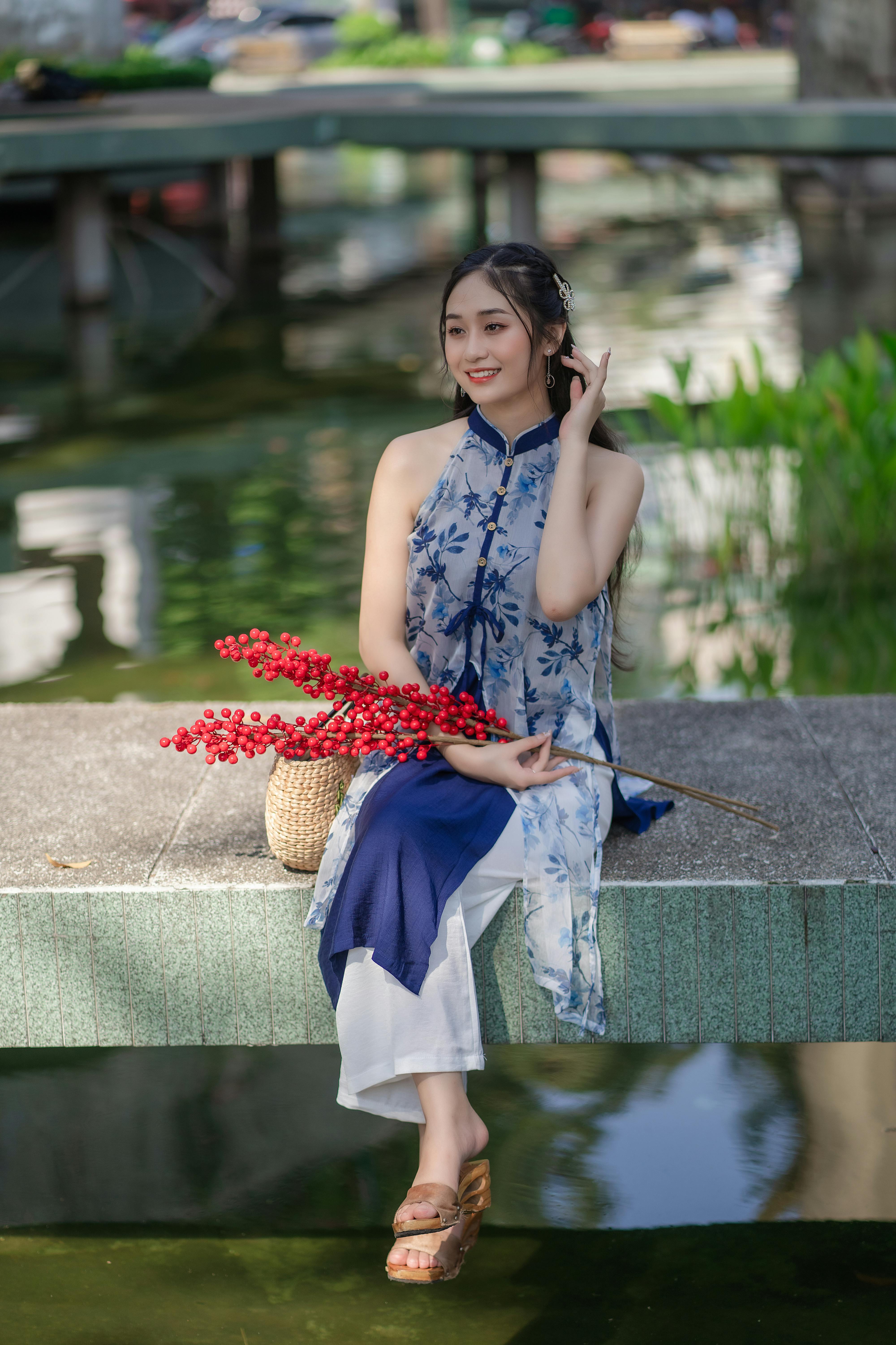 A young woman in a blue floral Vietnamese tunic sits by a pond, holding red flowers and smiling.