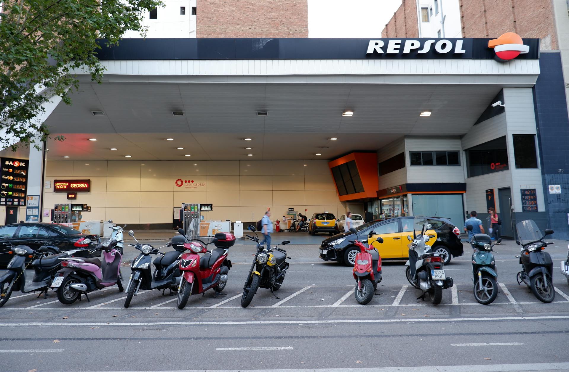 Repsol gas station in Barcelona, featuring parked motor scooters and taxis.