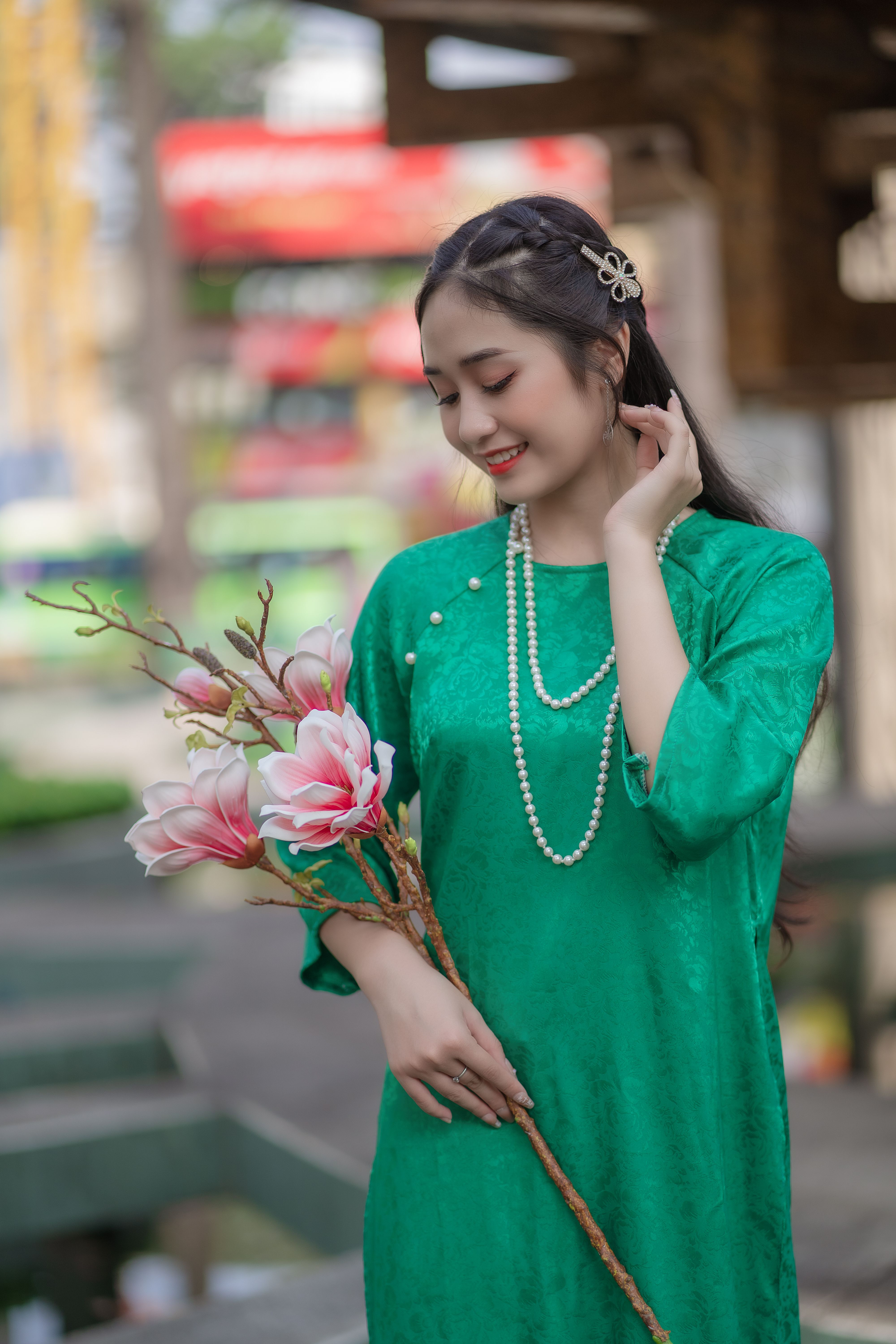 A young woman in an emerald dress gracefully holds pink flowers adorned with pearls outdoors.