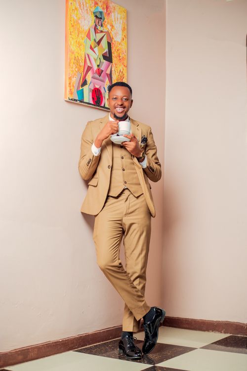 Free A fashionable man in a tan suit enjoying a cup of tea, smiling indoors with a vibrant painting. Stock Photo