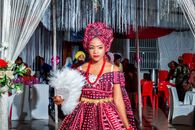 Beautiful woman in vibrant, traditional Nigerian attire holding a white fan during an indoor celebration.