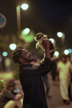 A young man joyfully lifts his fluffy puppy on a vibrant city street at night.