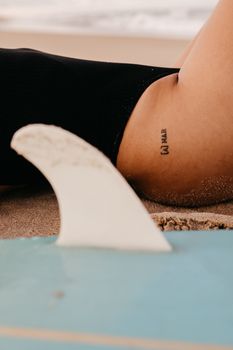 A woman relaxes on a sandy beach, featuring a surfboard and a visible tattoo.