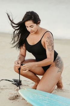 A woman in a swimming costume prepares her surfboard on a sunny beach, showcasing her tattoos.