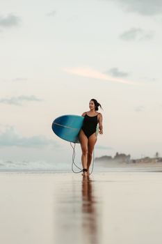 A woman in a swimsuit walks along the beach with her surfboard during sunset.