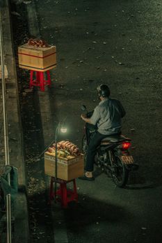 Motorcyclist pauses by street vendor stalls at night, dimly lit urban scene.