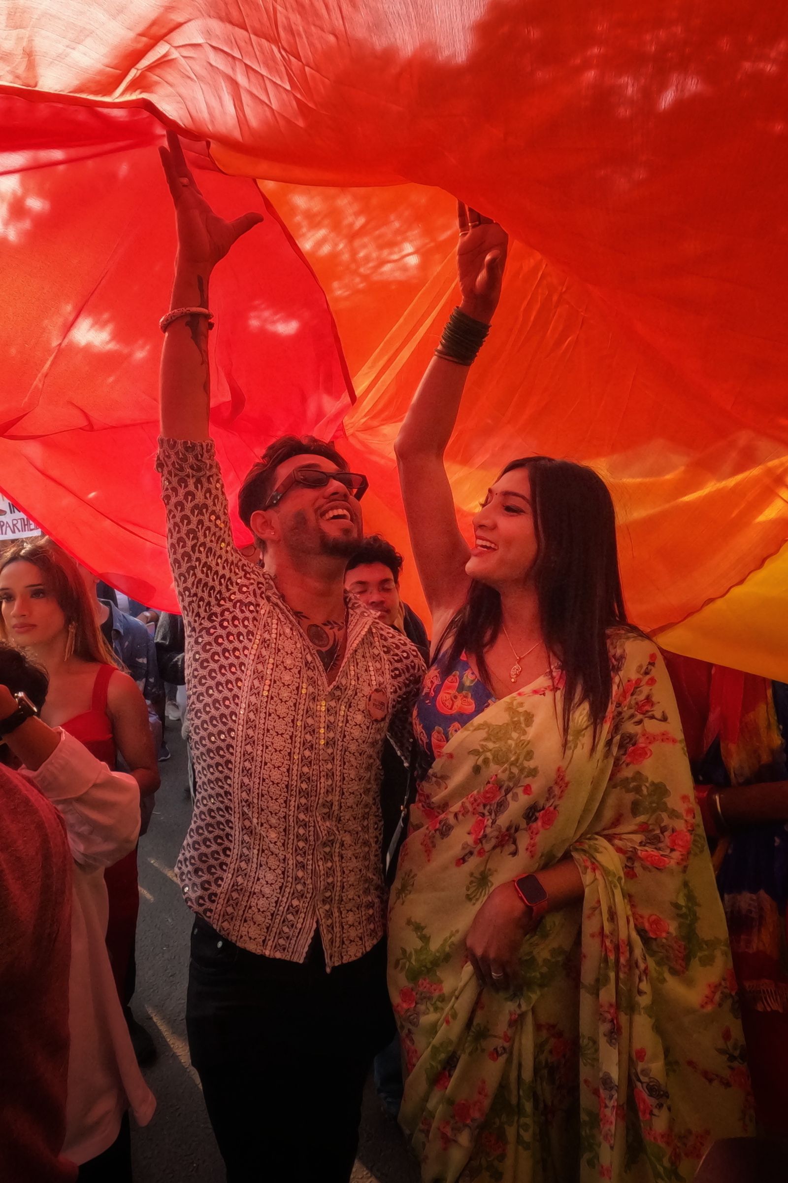 Two friends cheering under a giant rainbow banner at Kolkata Pride
