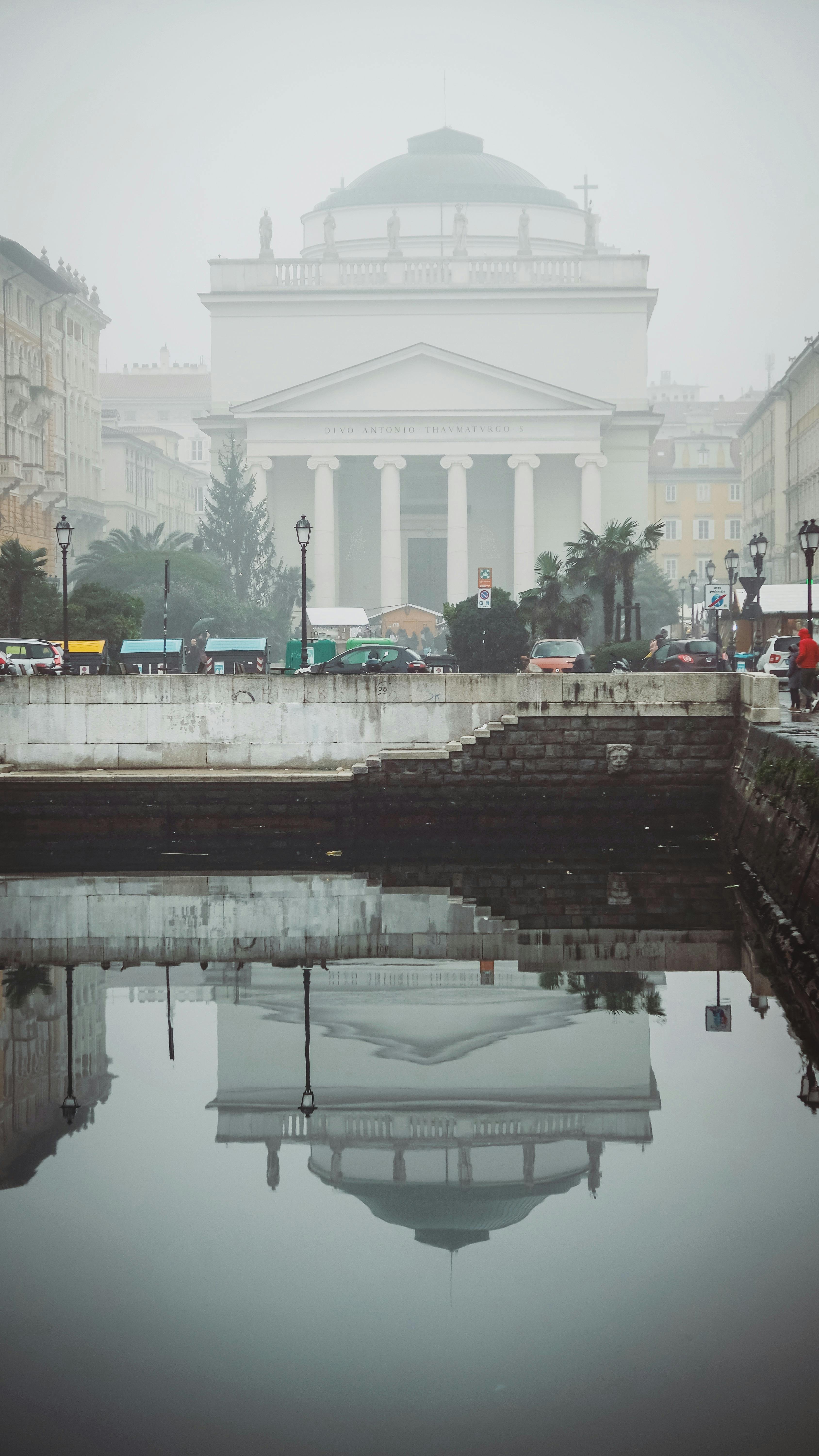 A historic white building with domed roof and columns reflected in a calm canal on a foggy day.