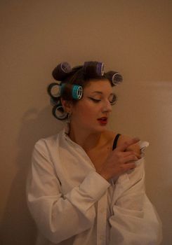 Woman in curlers and white shirt stands thoughtfully indoors with red lips.