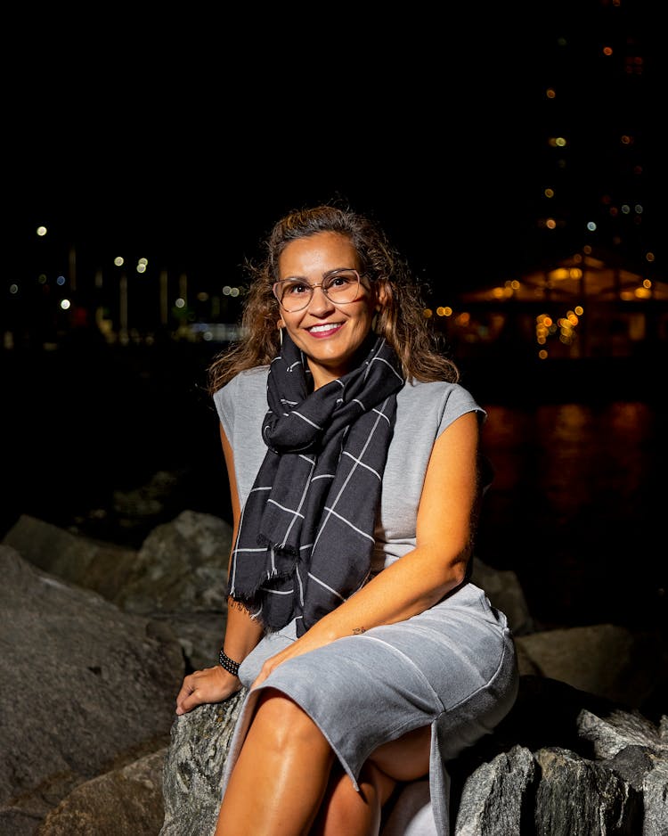 An Adult Woman Sitting On A Rock By The Water At Night And Smiling 