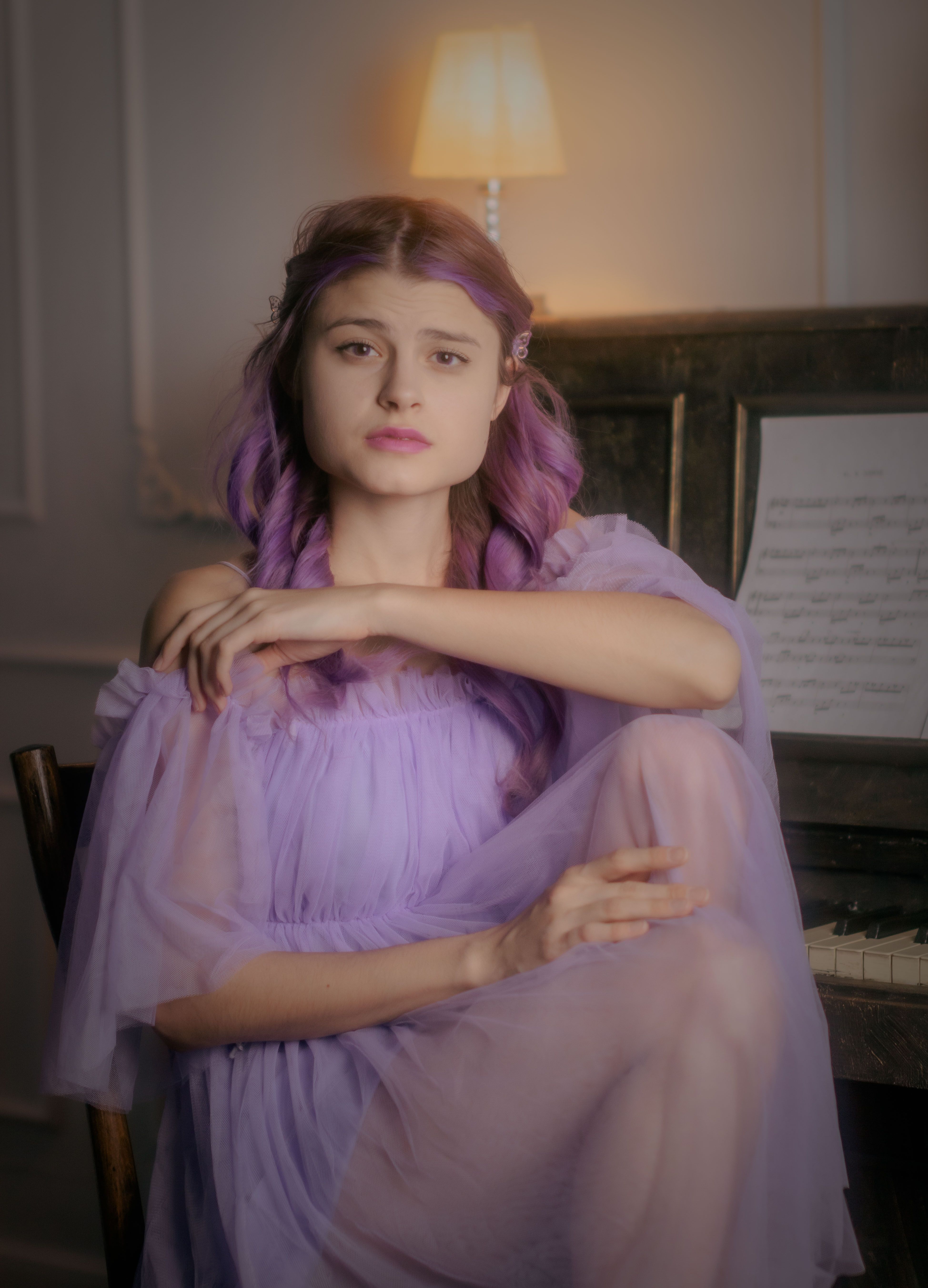 Young woman in lavender chiffon dress posing by a piano indoors.