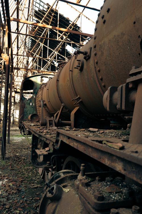 Free A rusted obsolete train inside a deteriorating industrial site, with sunlight filtering through broken roofs. Stock Photo