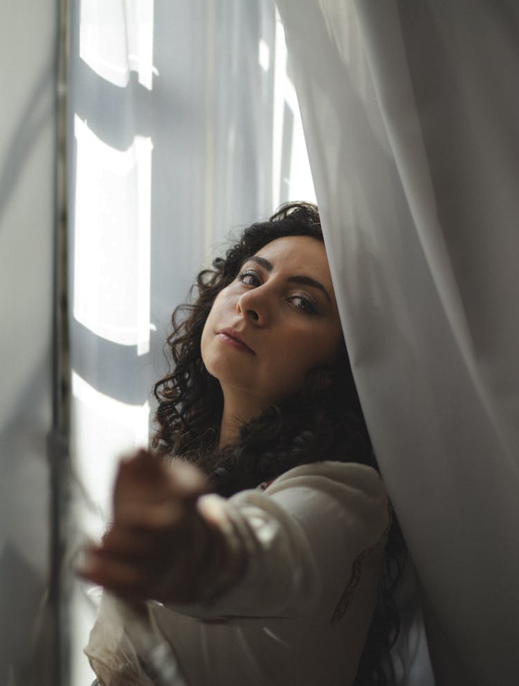 Young Woman With Curly Hair Standing Between The Curtains By The Window 