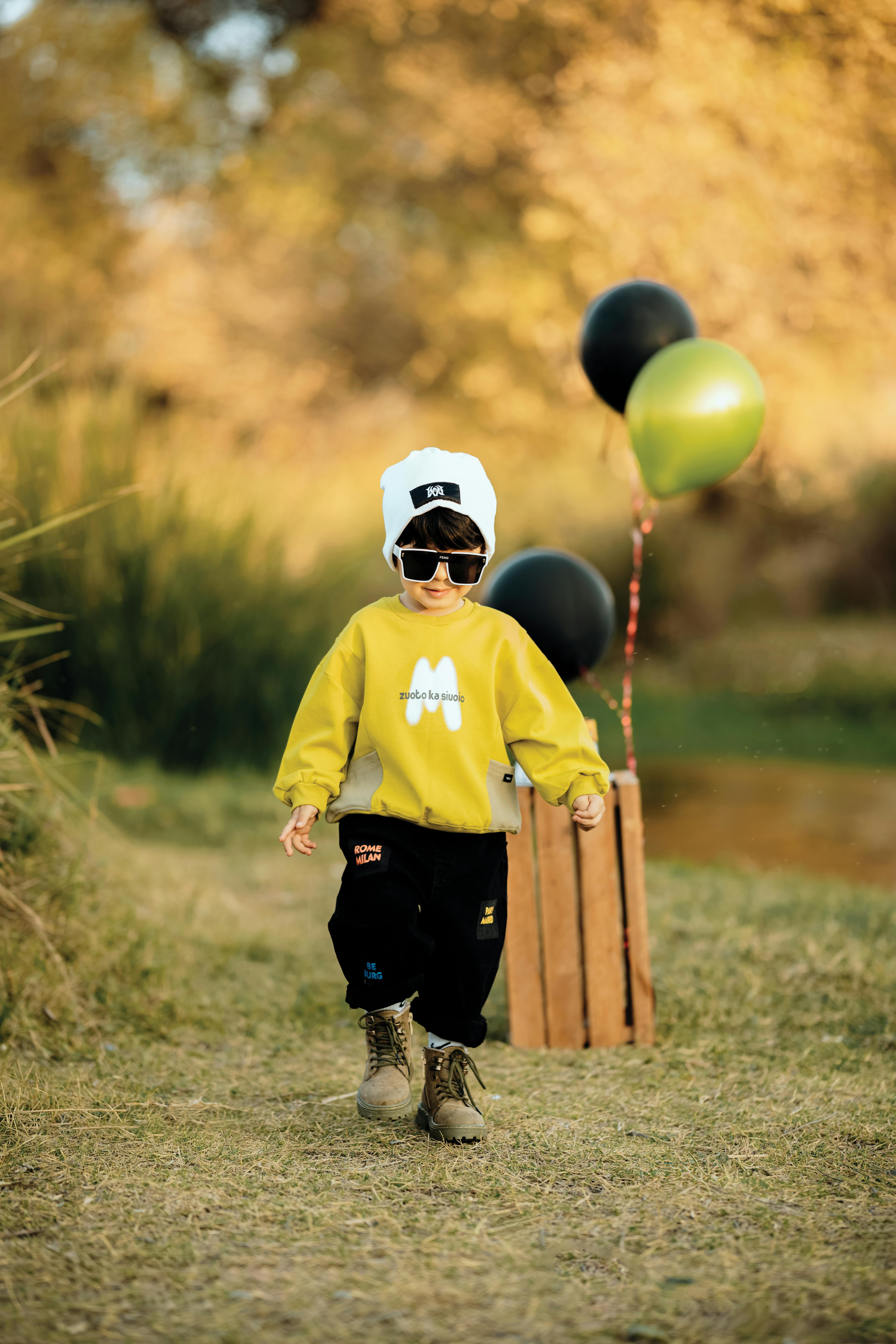 A trendy child wearing sunglasses walks through an autumn park with balloons and a wooden crate.