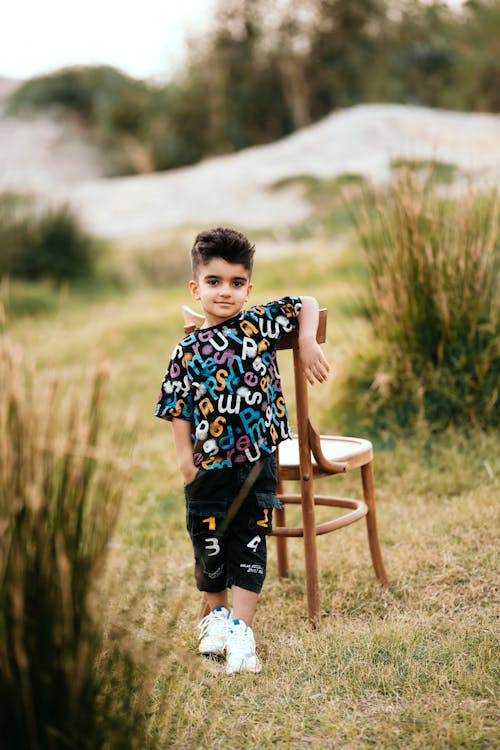 A Little Boy in a Fashionable Outfit Standing by the Chair Outside 
