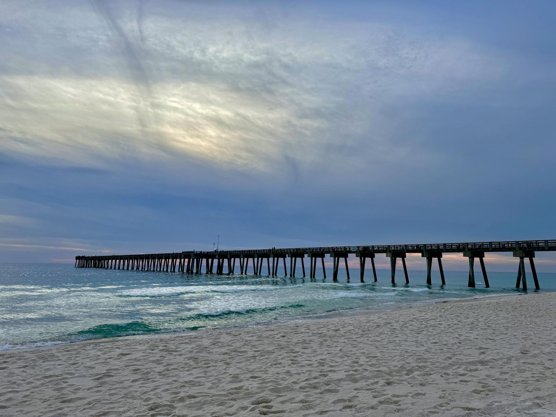 A tranquil beach scene with a long pier extending into the ocean at twilight, under a calming sky.