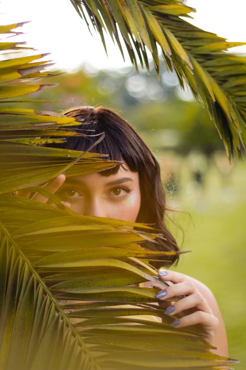 Free A woman peers through vibrant green palm leaves outdoors on a sunny day. Stock Photo